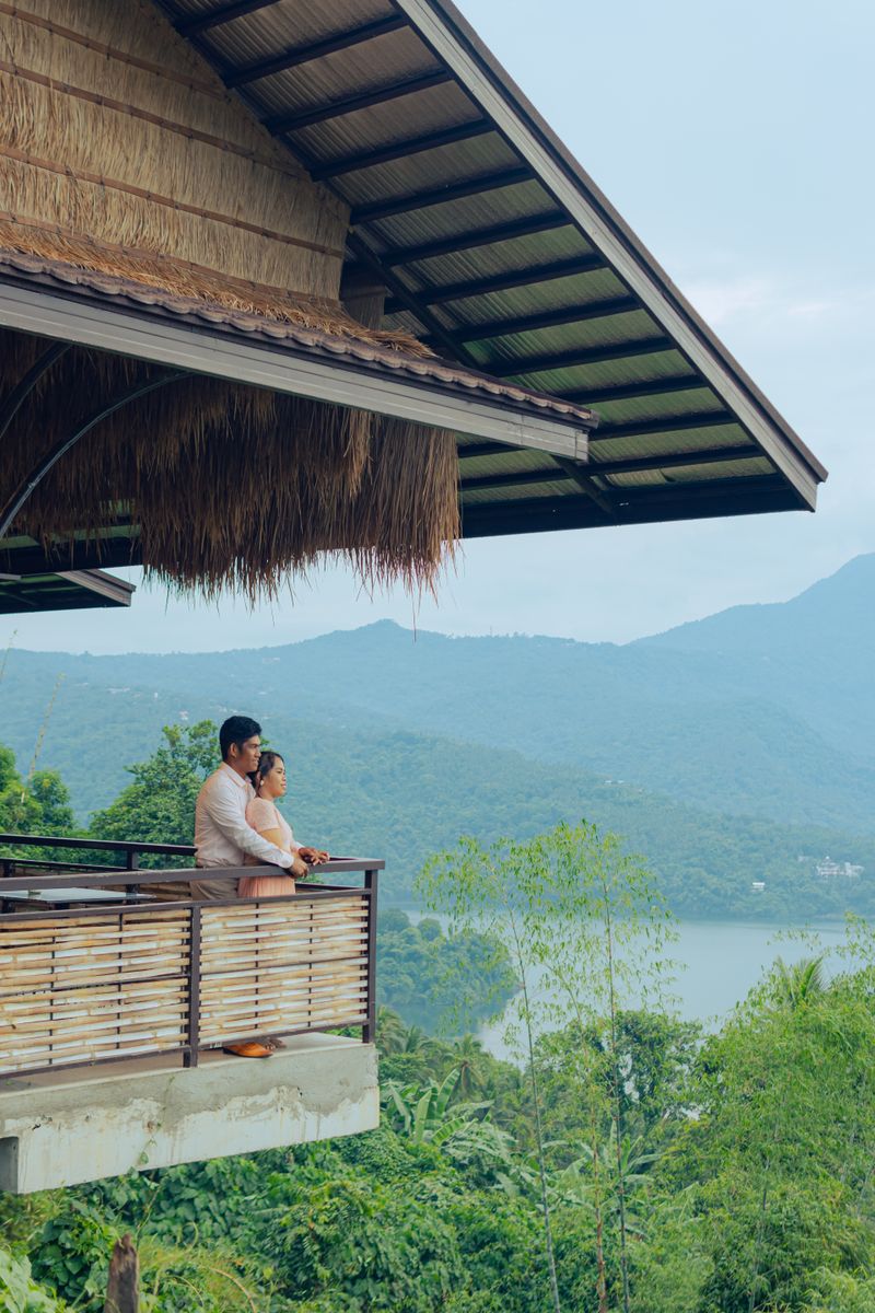 A couple stands on a balcony overlooking lush mountains and a tranquil lake.